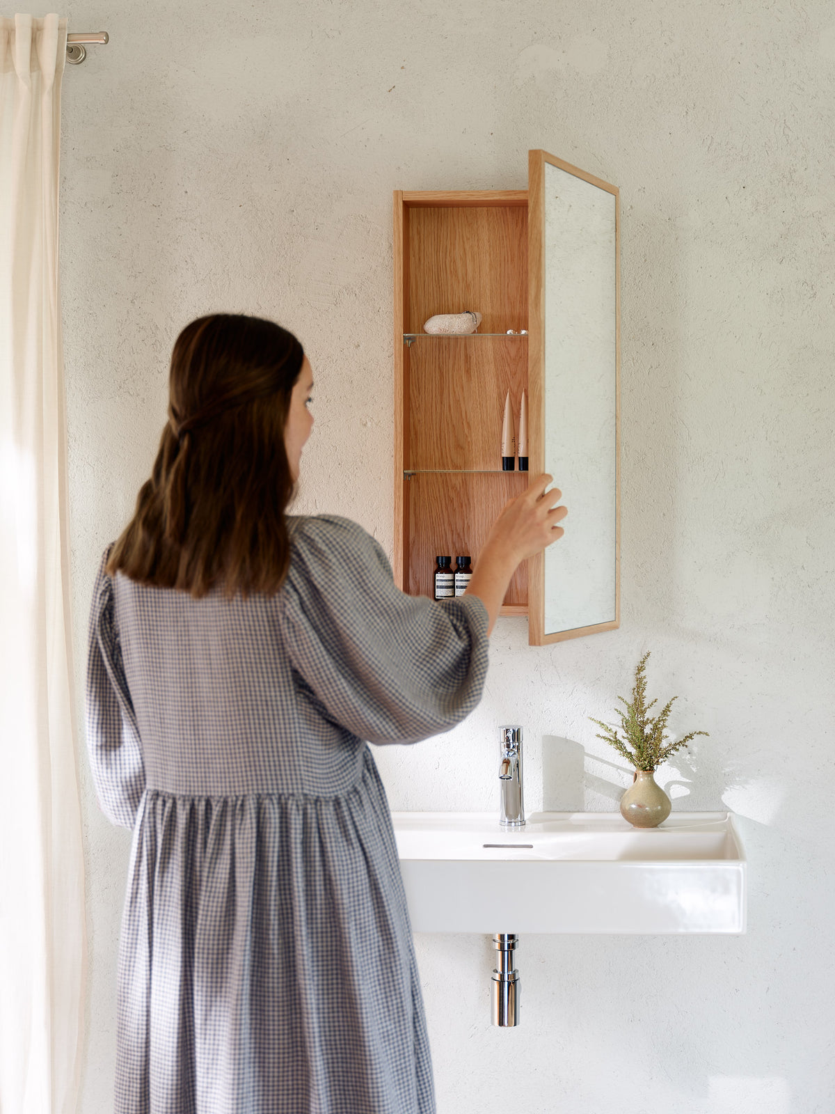 Woman opening a natural oak wall-mounted bathroom mirror cabinet with adjustable glass shelves
